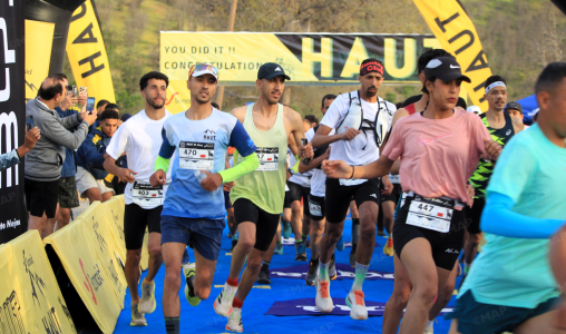 Group of runners sprint toward the finish on a blue carpet, bib numbers visible, with cheering spectators and yellow sponsor banners in the background.