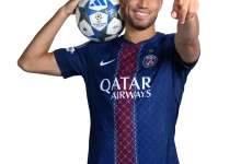 Soccer player in PSG jersey holds a Champions League ball over his shoulder and points toward the viewer, smiling confidently.