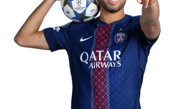 Soccer player in PSG jersey holds a Champions League ball over his shoulder and points toward the viewer, smiling confidently.