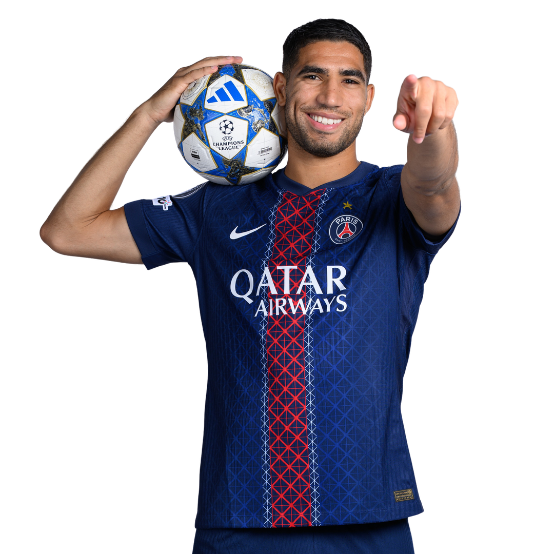 Soccer player in PSG jersey holds a Champions League ball over his shoulder and points toward the viewer, smiling confidently.