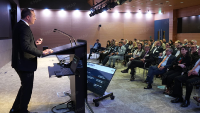 Male presenter in a suit speaks at a podium to an audience in a bright conference hall, with a laptop and screen nearby.