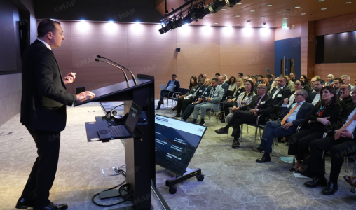 Male presenter in a suit speaks at a podium to an audience in a bright conference hall, with a laptop and screen nearby.