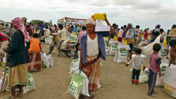 A crowded refugee aid distribution scene on a dusty ground: people carry large white bags and boxes, with aid staff and motorbikes in the background; a man in a tan skirt and white headscarf carries a yellow container on his shoulder.