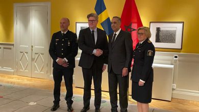 Four officials pose for a photo in a yellow-walled room; two men in suits shake hands while a police officer and a policewoman stand beside them, with blue-yellow and red flags in the background.