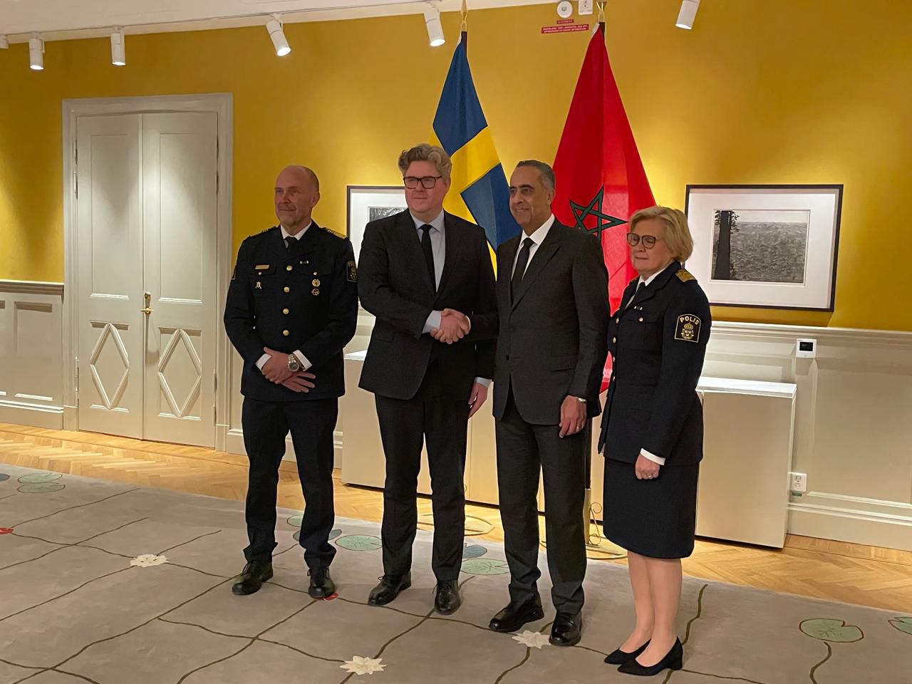 Four officials pose for a photo in a yellow-walled room; two men in suits shake hands while a police officer and a policewoman stand beside them, with blue-yellow and red flags in the background.