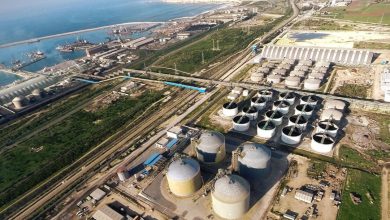 Aerial view of an industrial complex with numerous large cylindrical storage tanks near the coast, connected by rail lines and roads across a grassy landscape.