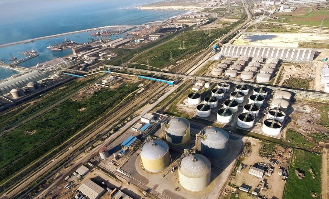 Aerial view of an industrial complex with numerous large cylindrical storage tanks near the coast, connected by rail lines and roads across a grassy landscape.