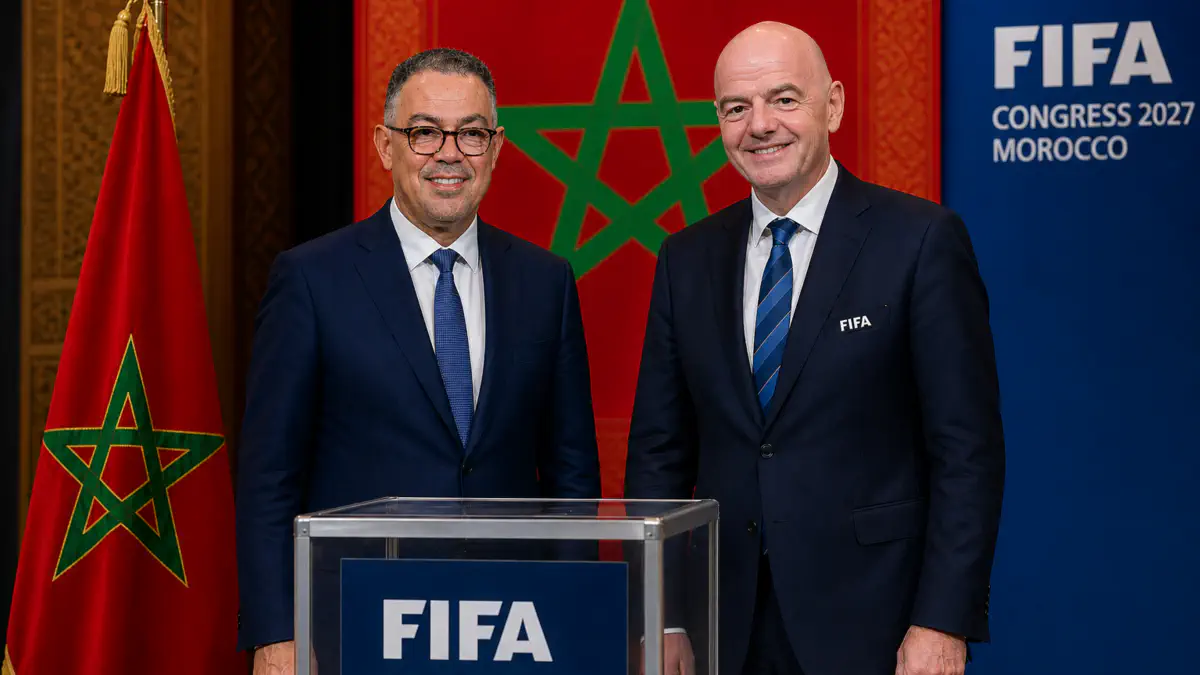 Two men in dark suits smile beside a transparent podium at a FIFA event, with the Moroccan flag and a FIFA Congress 2027 Morocco backdrop behind them.