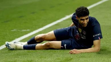 Soccer player in a dark kit lying on the grass, appearing injured near the sideline.