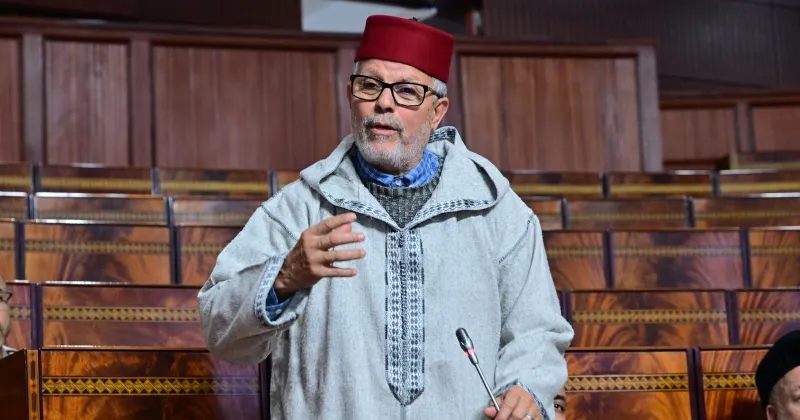 Man wearing a red fez and traditional gray robe speaks at a microphone in a wood-paneled legislative chamber.