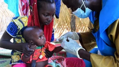 A healthcare worker in PPE gives a vaccination to a baby held by the mother outdoors under a thatched shelter.