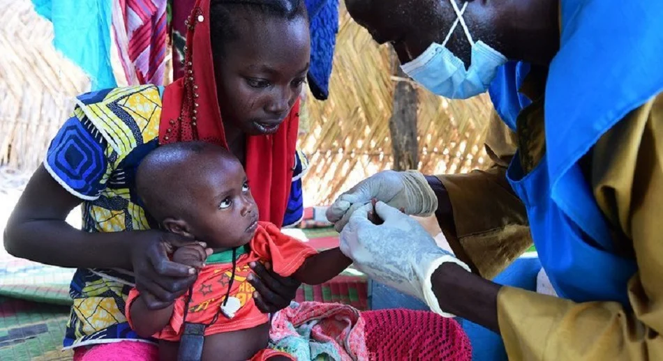 A healthcare worker in PPE gives a vaccination to a baby held by the mother outdoors under a thatched shelter.