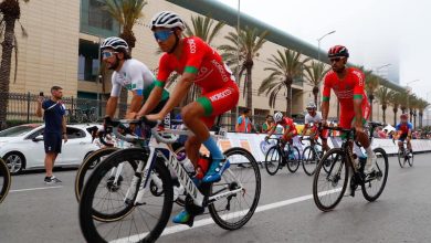Cyclists racing on a city street, wearing team kits and helmets, with spectators and palm trees in the background.