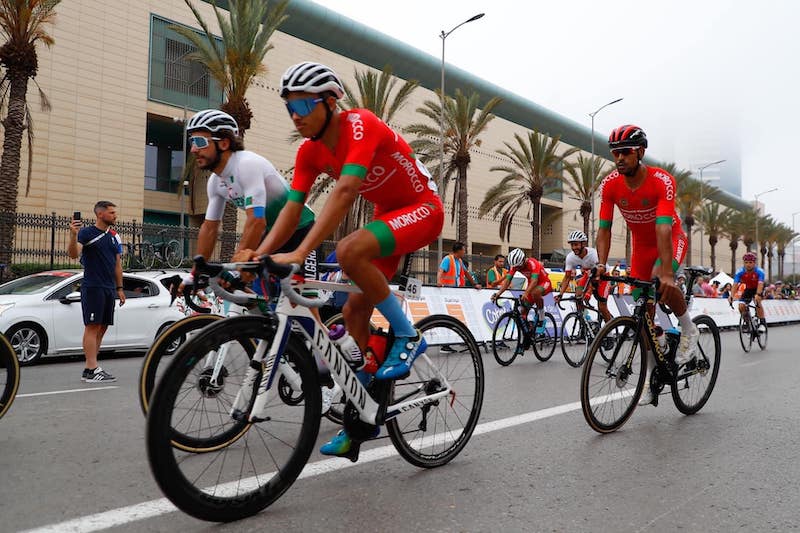 Cyclists racing on a city street, wearing team kits and helmets, with spectators and palm trees in the background.