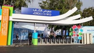 Entrance to a book fair with a blue Arabic sign, curved white canopies, colorful pillars, and turnstiles at the gate.