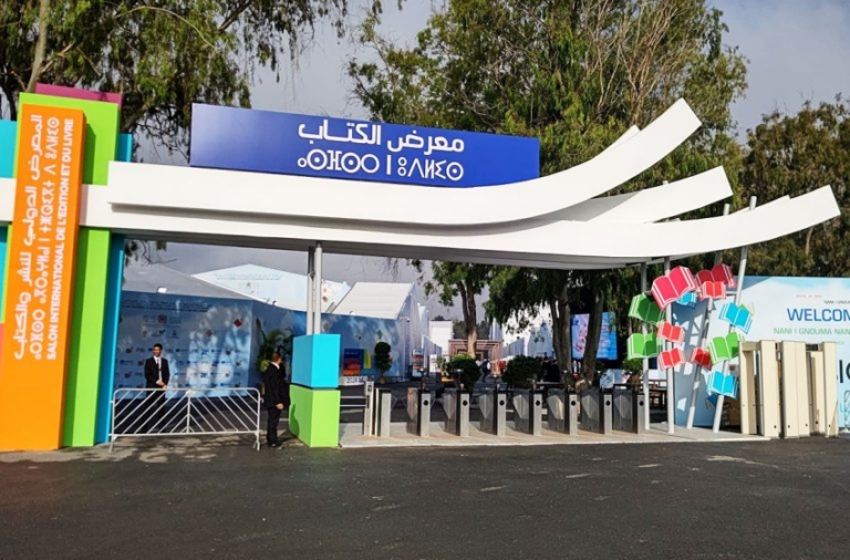 Entrance to a book fair with a blue Arabic sign, curved white canopies, colorful pillars, and turnstiles at the gate.
