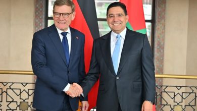 Two men in dark suits shake hands and smile, with German and Jordanian flags in the background behind them.