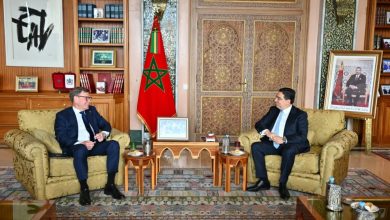Two men in suits sit on ornate armchairs facing each other in a formal office with a Moroccan flag between them.