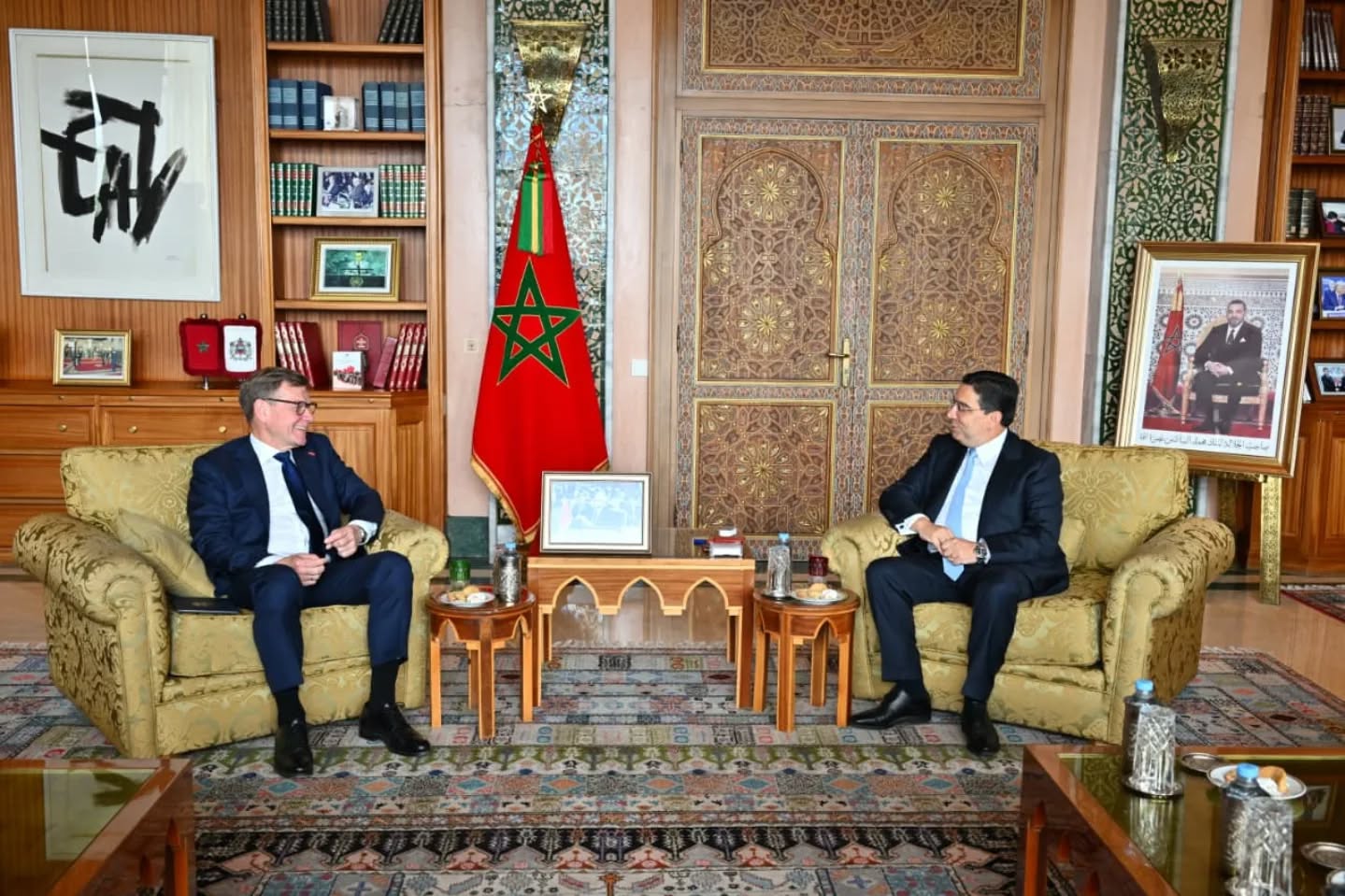 Two men in suits sit on ornate armchairs facing each other in a formal office with a Moroccan flag between them.