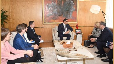 Diplomatic/business meeting in a wood-paneled room; a man in a dark suit speaks to five colleagues seated around a coffee table.