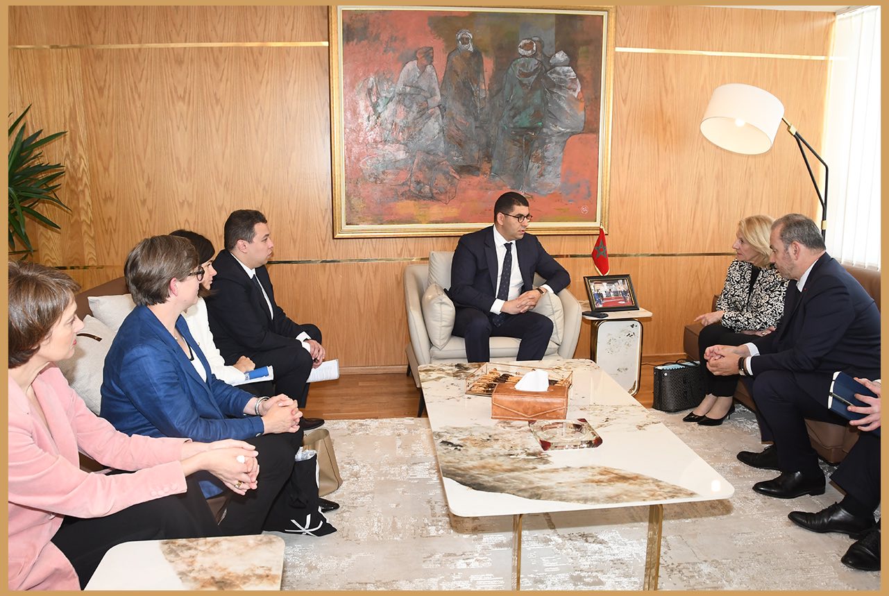 Diplomatic/business meeting in a wood-paneled room; a man in a dark suit speaks to five colleagues seated around a coffee table.