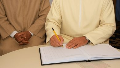 Person in pale robes signs an open ledger on a round table while another person sits beside with hands folded.