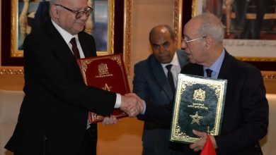 Two men in suits shake hands while each holds an ornate certificate folder, with a small Moroccan flag visible in the foreground.