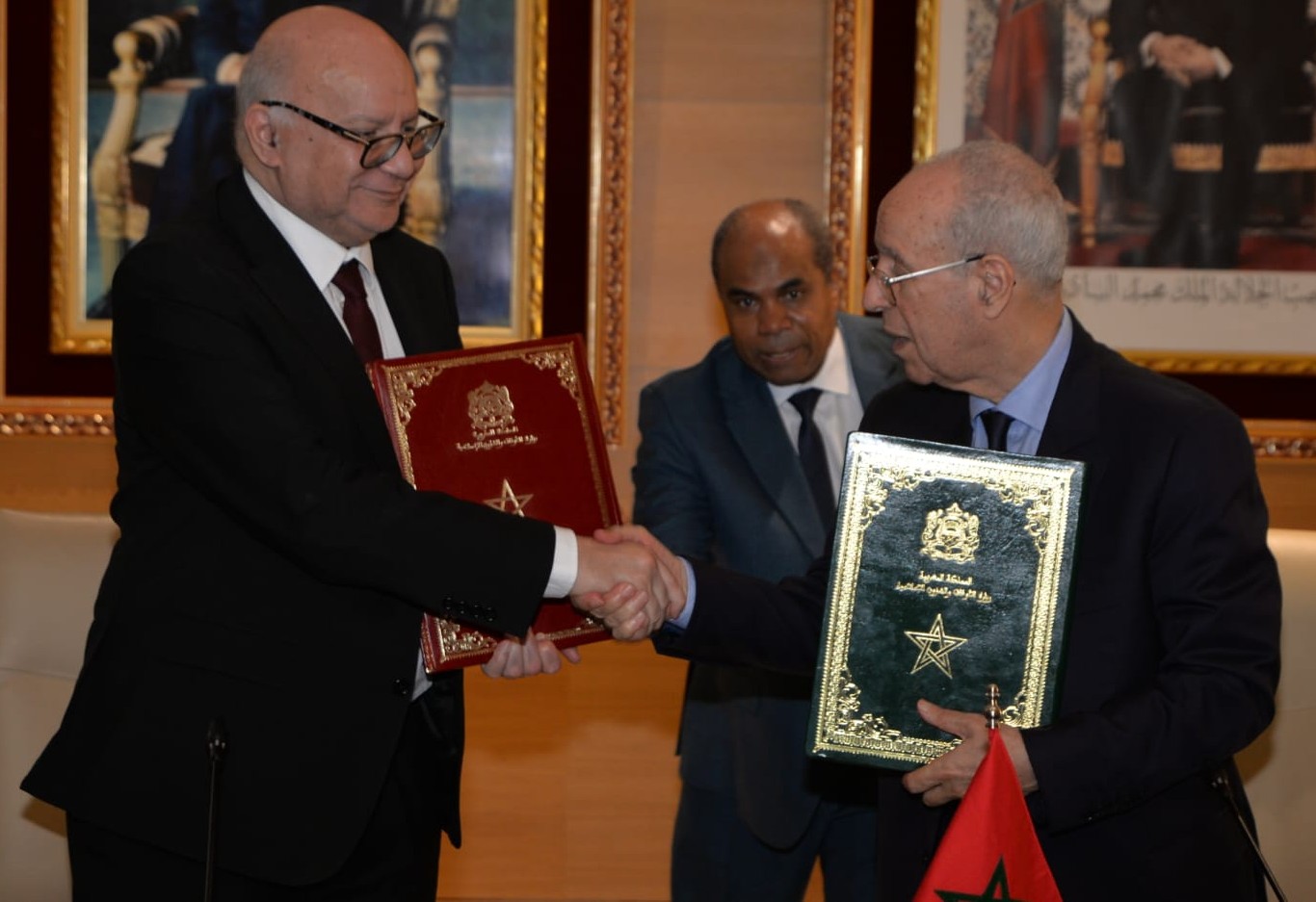 Two men in suits shake hands while each holds an ornate certificate folder, with a small Moroccan flag visible in the foreground.