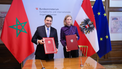 Two officials sign agreement documents at a formal ceremony, flanked by the Moroccan flag on the left, Austrian flag, and EU flag in a government meeting room.