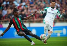 Two soccer players contest the ball on a grassy pitch during a match, one in black with red and green stripes and the other in white and green.