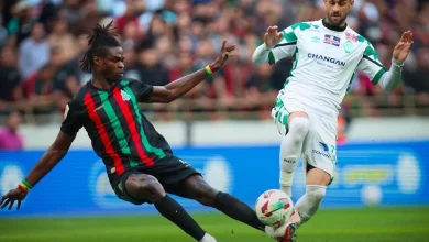 Two soccer players contest the ball on a grassy pitch during a match, one in black with red and green stripes and the other in white and green.