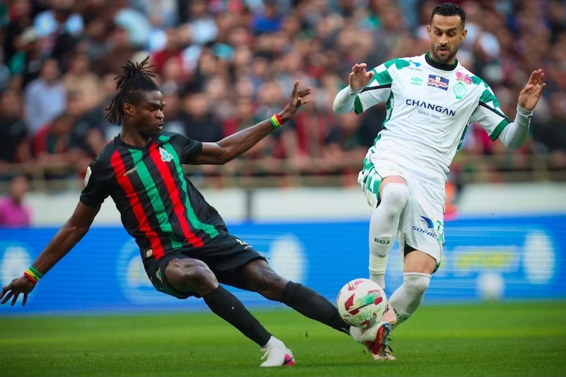 Two soccer players contest the ball on a grassy pitch during a match, one in black with red and green stripes and the other in white and green.