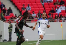 Soccer match moment: a Black player in a dark jersey challenges a white-clad player near the sideline with red stadium seats in the background.