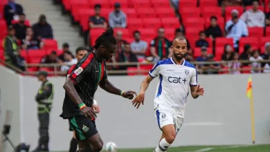 Soccer match moment: a Black player in a dark jersey challenges a white-clad player near the sideline with red stadium seats in the background.