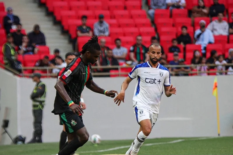 Soccer match moment: a Black player in a dark jersey challenges a white-clad player near the sideline with red stadium seats in the background.