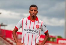 Soccer player posing with hands on hips in a white jersey with red stripes on a football pitch.