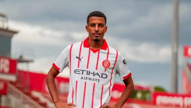 Soccer player posing with hands on hips in a white jersey with red stripes on a football pitch.