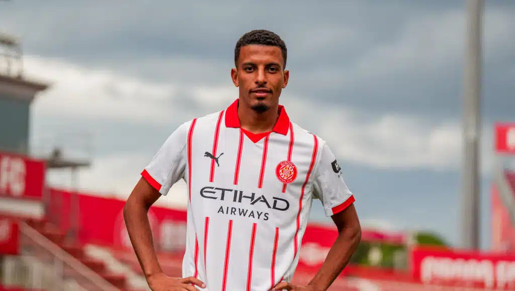 Soccer player posing with hands on hips in a white jersey with red stripes on a football pitch.