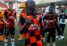 Soccer players in red-and-black checkered jerseys celebrate on the field; foreground player pumps fists in jubilation as teammates cheer behind.