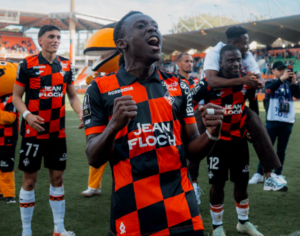 Soccer players in red-and-black checkered jerseys celebrate on the field; foreground player pumps fists in jubilation as teammates cheer behind.