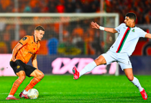 Soccer player in orange controls the ball as a white-jersey opponent reaches to intercept near the goal.