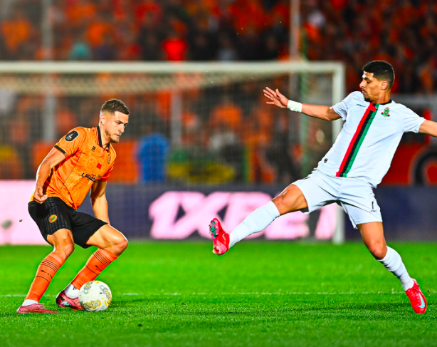 Soccer player in orange controls the ball as a white-jersey opponent reaches to intercept near the goal.