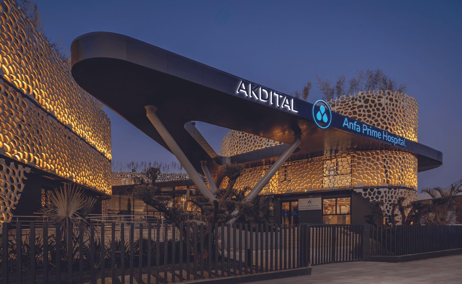 Modern hospital exterior at dusk with a curved canopy and illuminated AKDITAL Anfa Prime Hospital sign against a honeycomb-lit facade