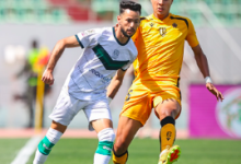 Two soccer players duel for the ball on a sunny green field; one wears white with green accents, the other yellow.