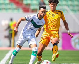 Two soccer players duel for the ball on a sunny green field; one wears white with green accents, the other yellow.
