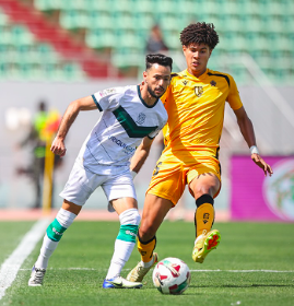 Two soccer players duel for the ball on a sunny green field; one wears white with green accents, the other yellow.