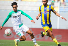Soccer match scene with a player in a green‑and‑white kit dribbling the ball while a blue‑and‑yellow striped opponent chases him.