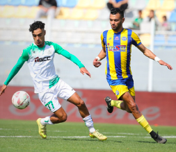 Soccer match scene with a player in a green‑and‑white kit dribbling the ball while a blue‑and‑yellow striped opponent chases him.