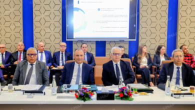 Formal business meeting with executives seated at a long table, a presentation screen behind them showing slides.