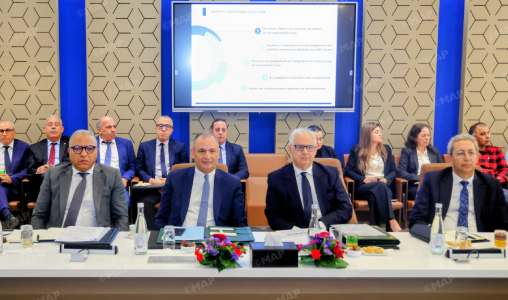 Formal business meeting with executives seated at a long table, a presentation screen behind them showing slides.